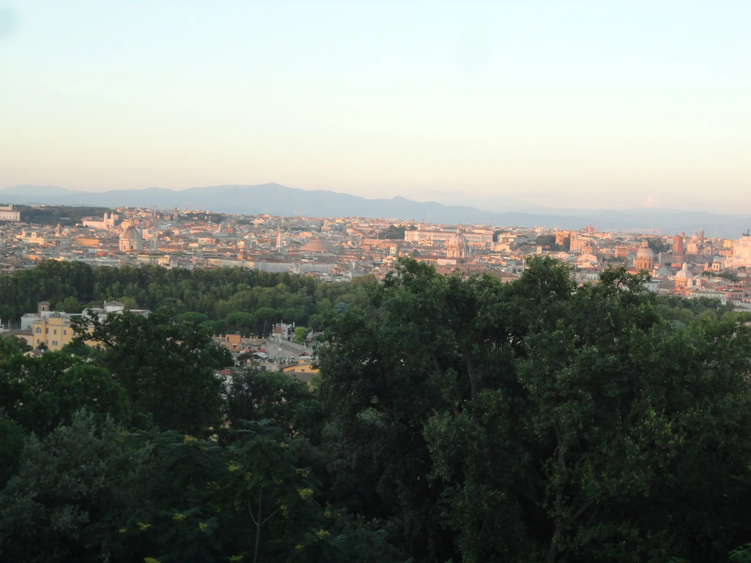 Rome from the Gianicolo in the first light of day: domes and rooftops under a pale peach haze, with the Alban Hills behind.