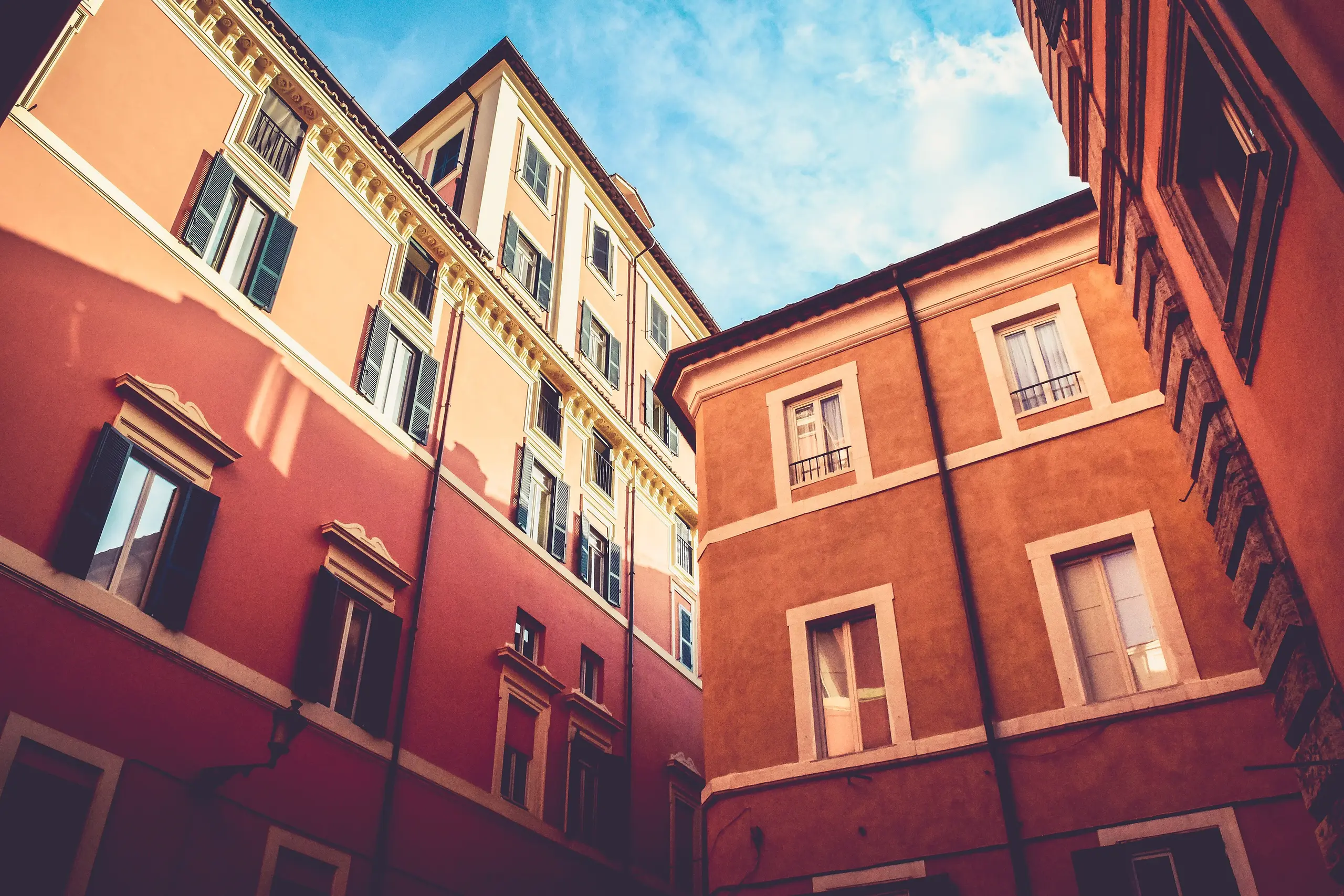 Looking up at ochre and terracotta palazzo facades with shuttered windows against a blue Roman sky.