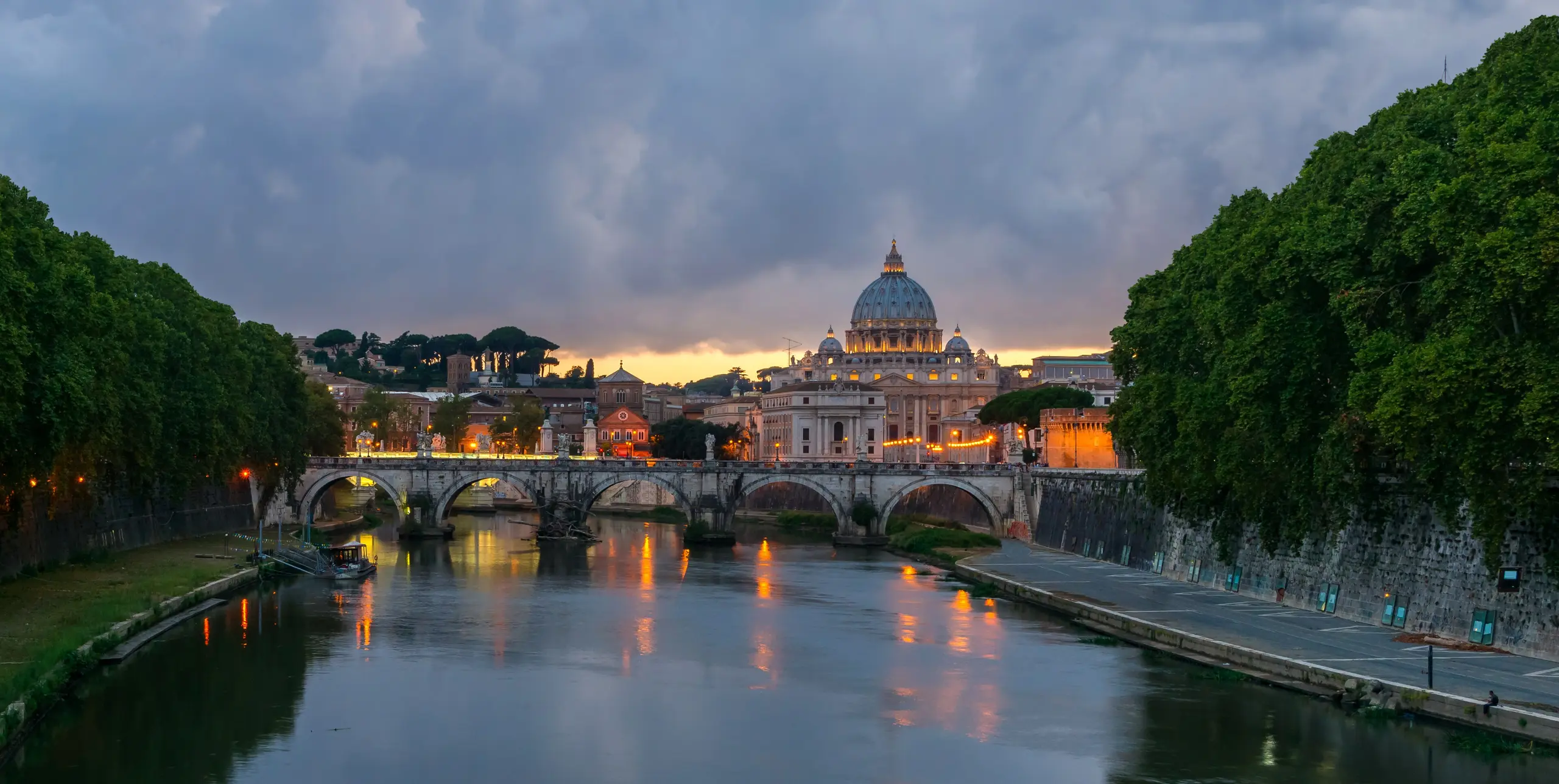 The Ponte Sant'Angelo's stone arches over the Tiber at dusk, lamps lit and the sky fading to blue.
